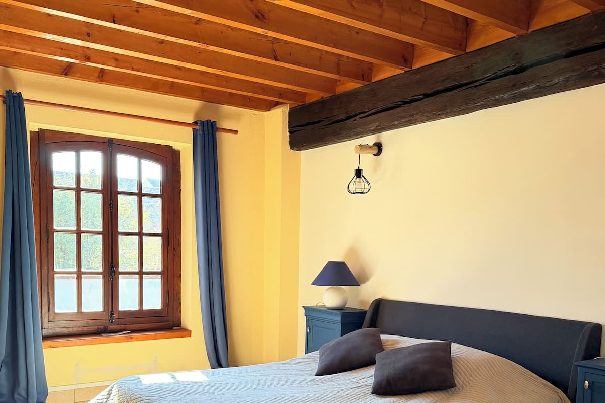 Een slaapkamer met een bed, een nachtkastje, een lamp, een raam met gordijnen en een houten plafond.