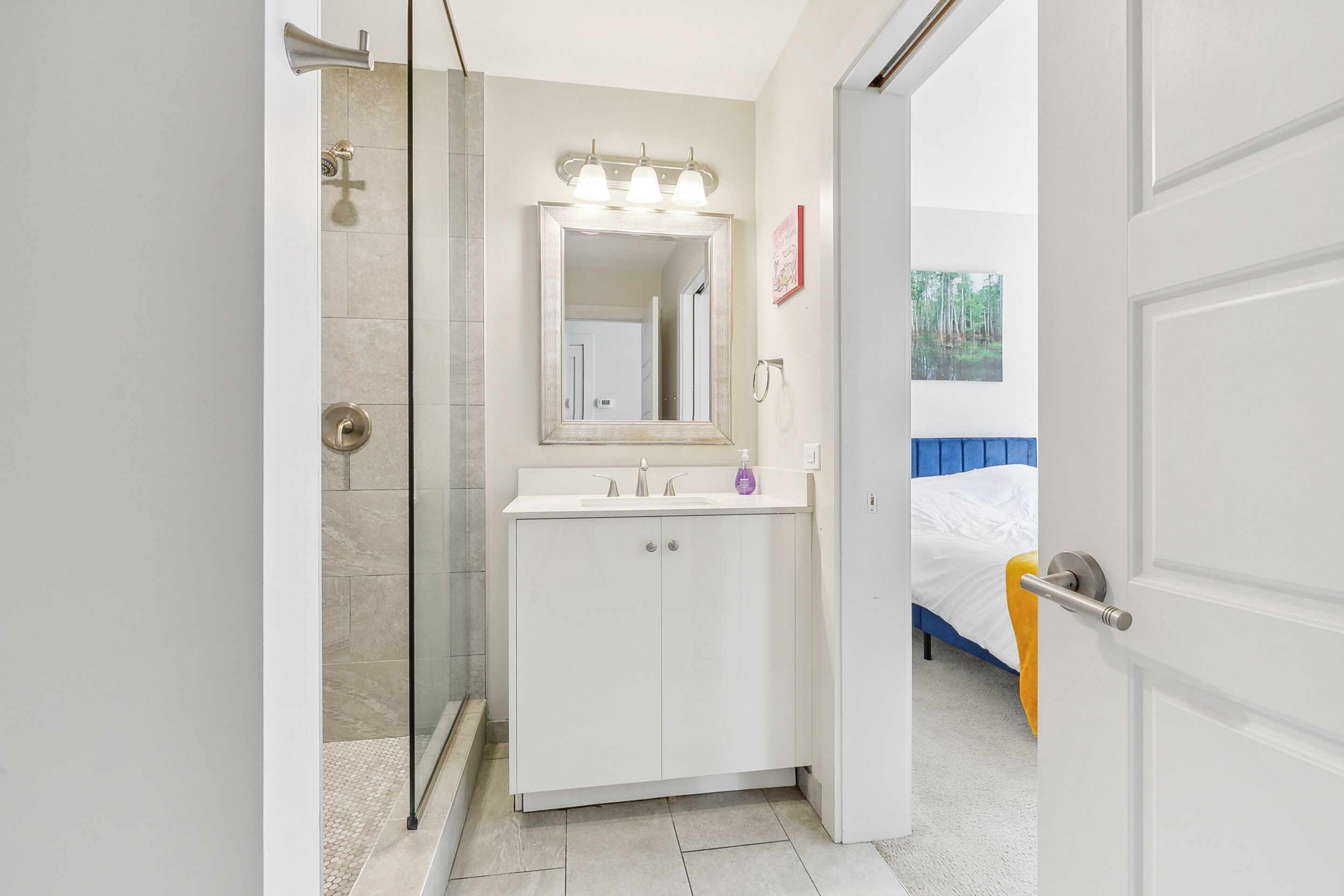 A bathroom with a shower, a sink with a mirror, and a bed in the adjacent room.