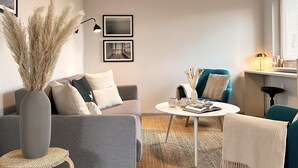 Ett modernt vardagsrum med en grå soffa, en turkos fåtölj och ett runt soffbord. På väggen hänger två inramade tavlor, och det står en hög vas med pampasgräs samt en lampa på sidobordet.