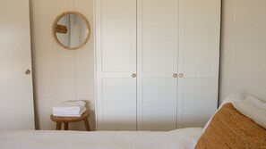 A bedroom with a bed, a wooden stool with folded towels, a round mirror, and a white wardrobe with two doors. - The Mullet - With Water Views (Nambucca Heads)