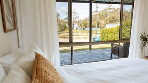 A bedroom with a large window, a bed with white linens, and a view of a paved area with greenery and a building. - The Mullet - With Water Views (Nambucca Heads)