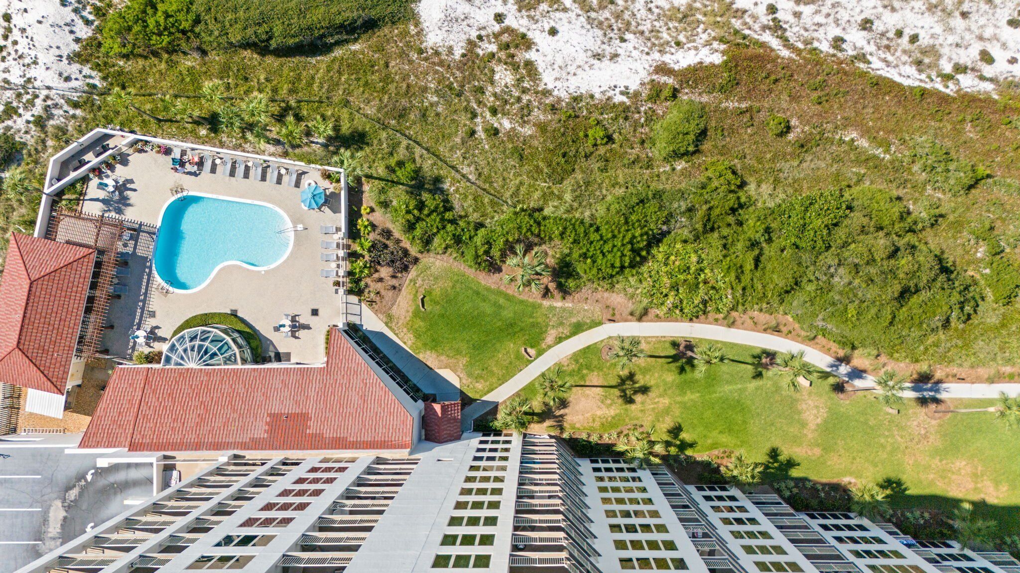 An aerial view of a hotel with a swimming pool, a rooftop terrace, and a curved pathway leading to a green area with trees.