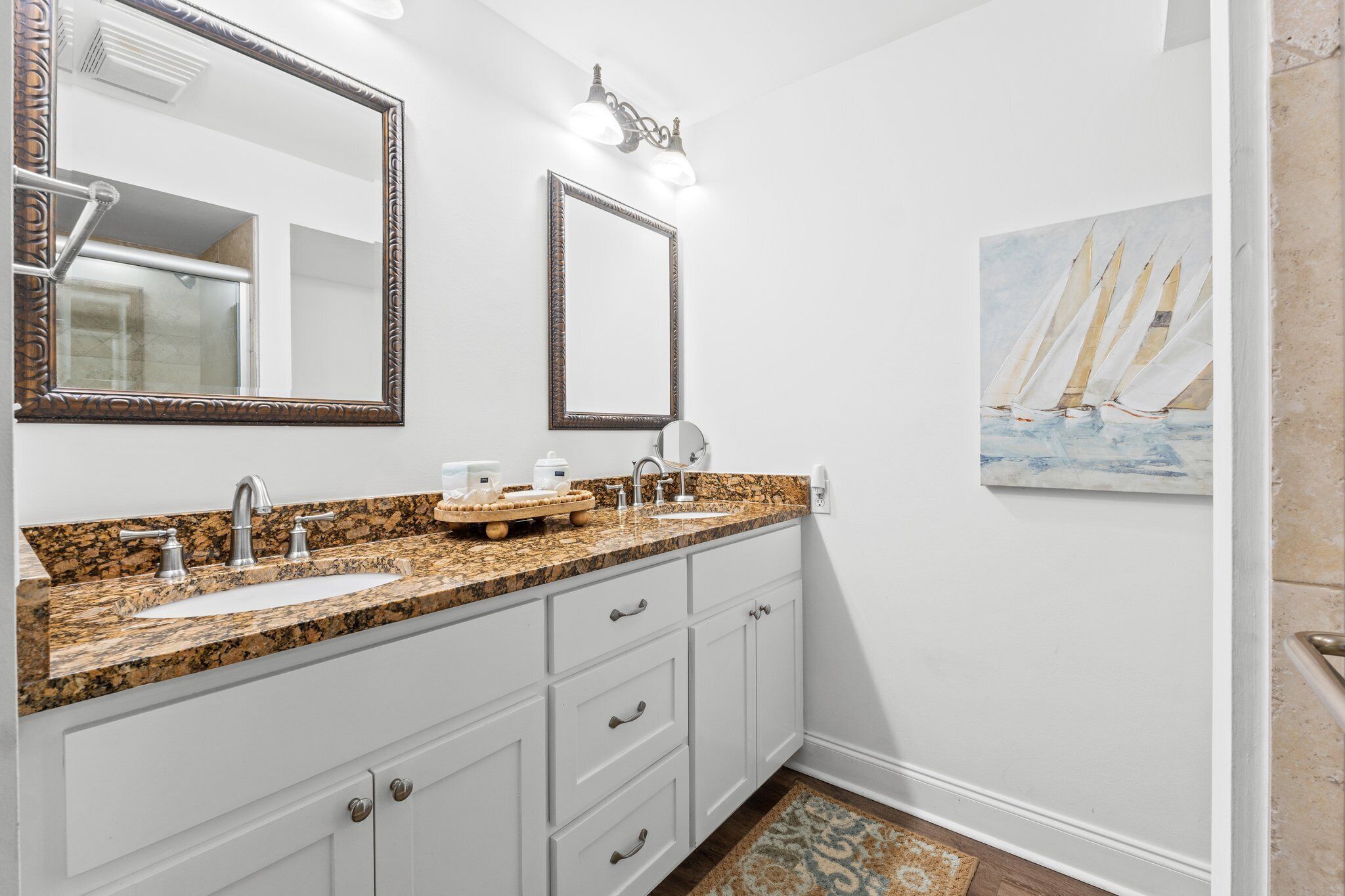 A bathroom with a granite countertop, two sinks, a large mirror, and a painting of sailboats.