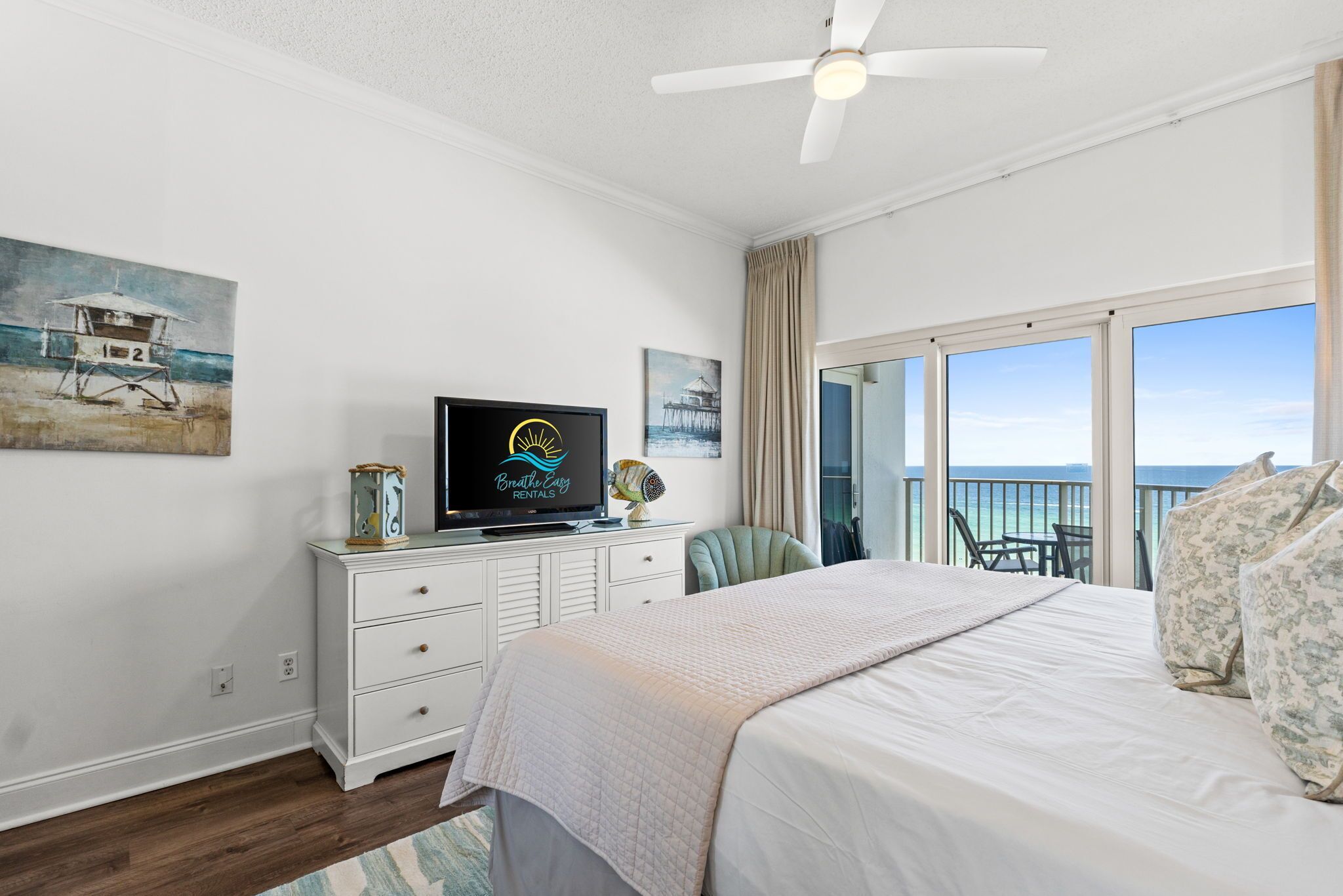 A bedroom with a bed, a television, a desk with a computer, and a balcony with a view of the ocean.