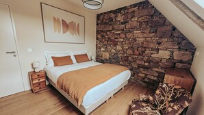 A bedroom with a stone wall, a bed with a brown blanket, a nightstand, and a chair with floral upholstery. - Le Lodge #1 with garden and pool access - Perros Guirec (Perros-Guirec)