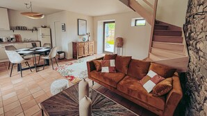 A modern living room with a brown sofa, a dining area with a table and chairs, a kitchen with a refrigerator, and a staircase. - Le Lodge #1 with garden and pool access - Perros Guirec (Perros-Guirec)