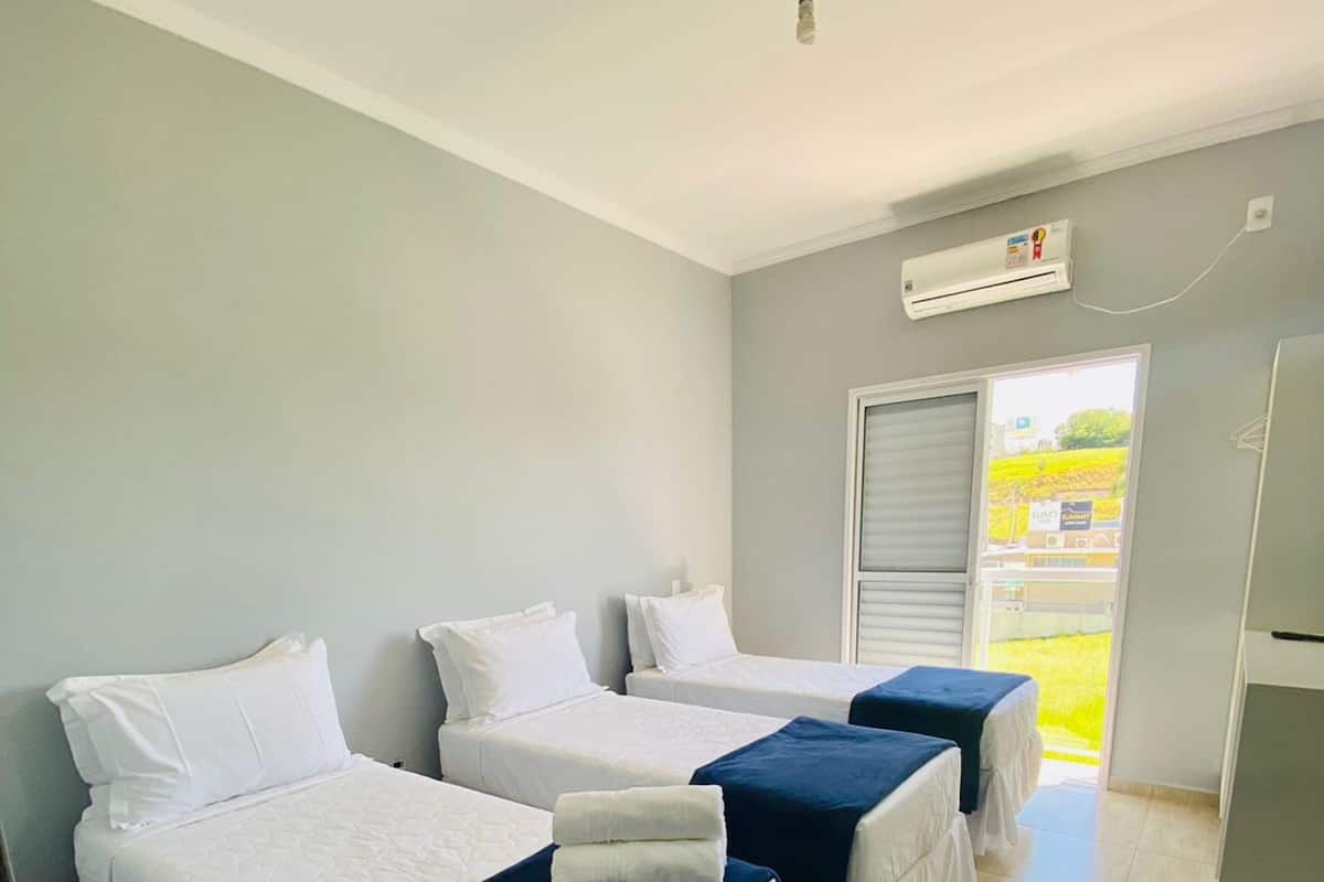 Ein Hotelzimmer mit drei Einzelbetten, jedes mit weißer Bettwäsche und einem blauen Akzent. Es gibt einen Deckenventilator, eine Klimaanlage und ein Fenster mit Blick ins Freie.