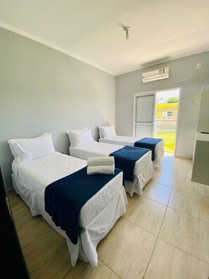 Ein Hotelzimmer mit drei Einzelbetten, jedes mit weißer Bettwäsche und einem blauen Akzent. Es gibt einen Deckenventilator, eine Klimaanlage und ein Fenster mit Blick ins Freie.