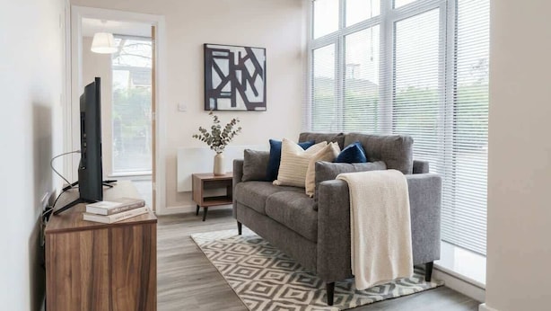 A modern living room with a grey sofa, a wooden side table, a flat-screen TV, and a large window with blinds. - Manchester & Salford | 1BED Apartment with Parking (Salford)