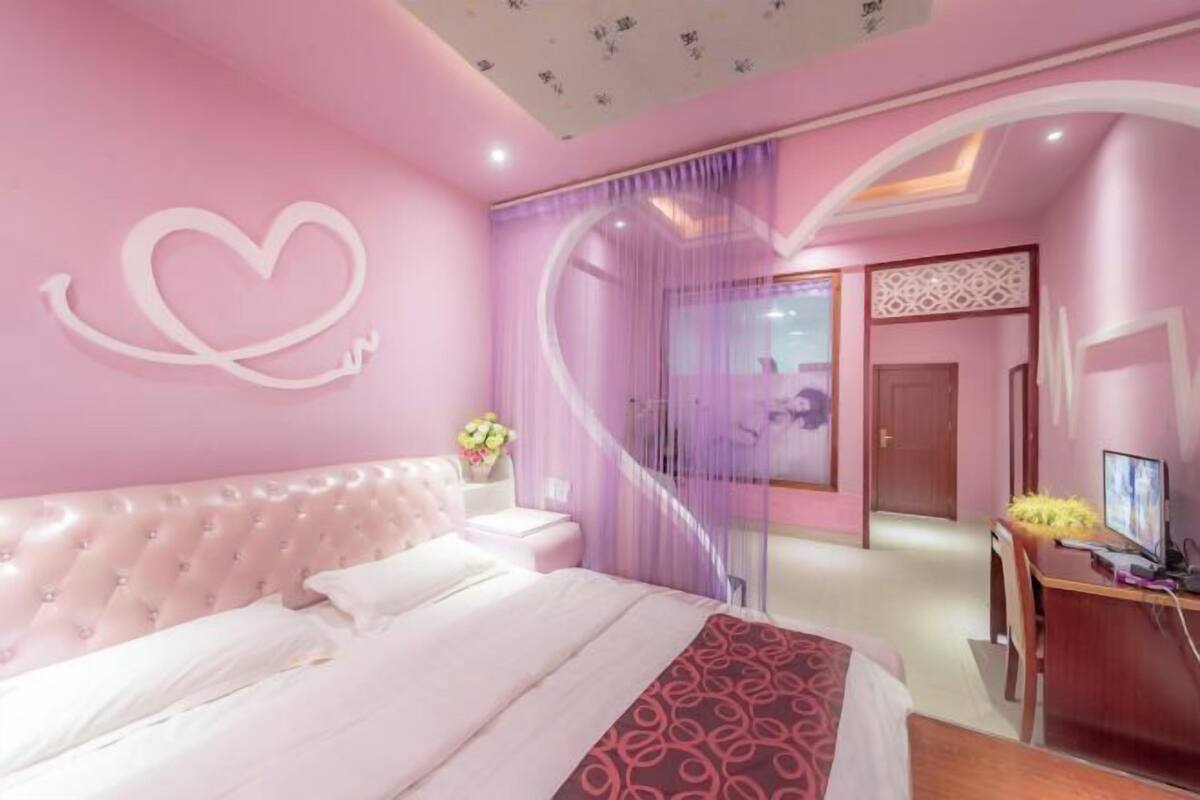 Een roze slaapkamer met een gestoffeerd hoofdeinde, een bed met witte beddengoed, een bureau met een computer en een decoratief hart aan de muur.