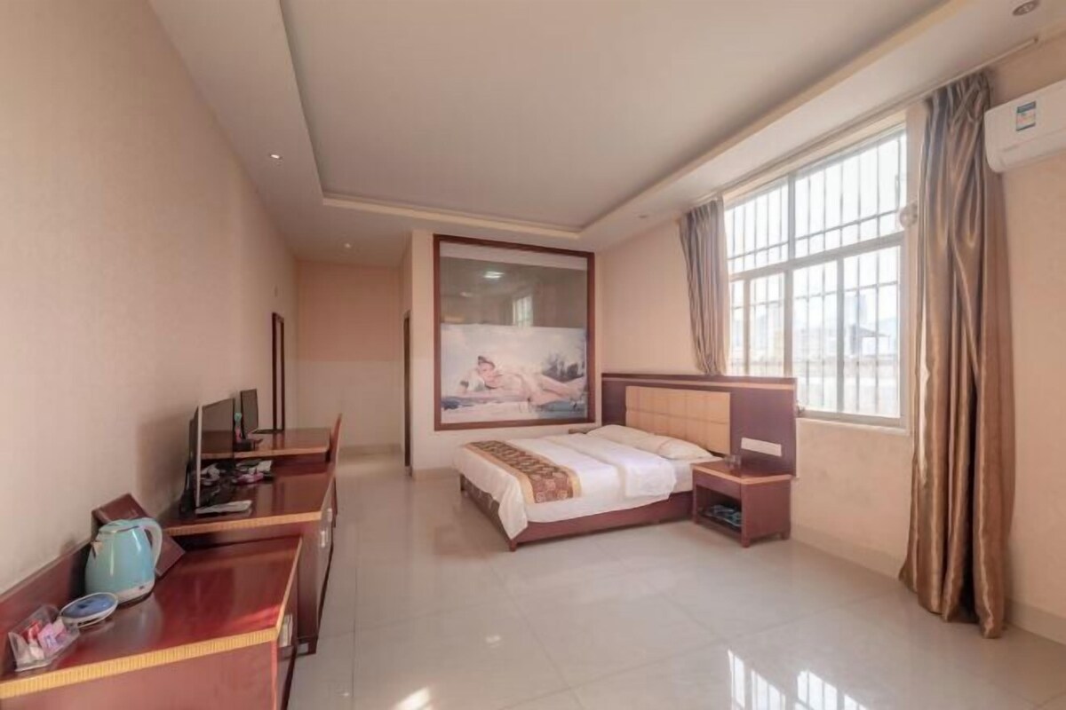 A hotel room with a bed, bedside tables, a desk with a kettle, a TV, and a large window with curtains.