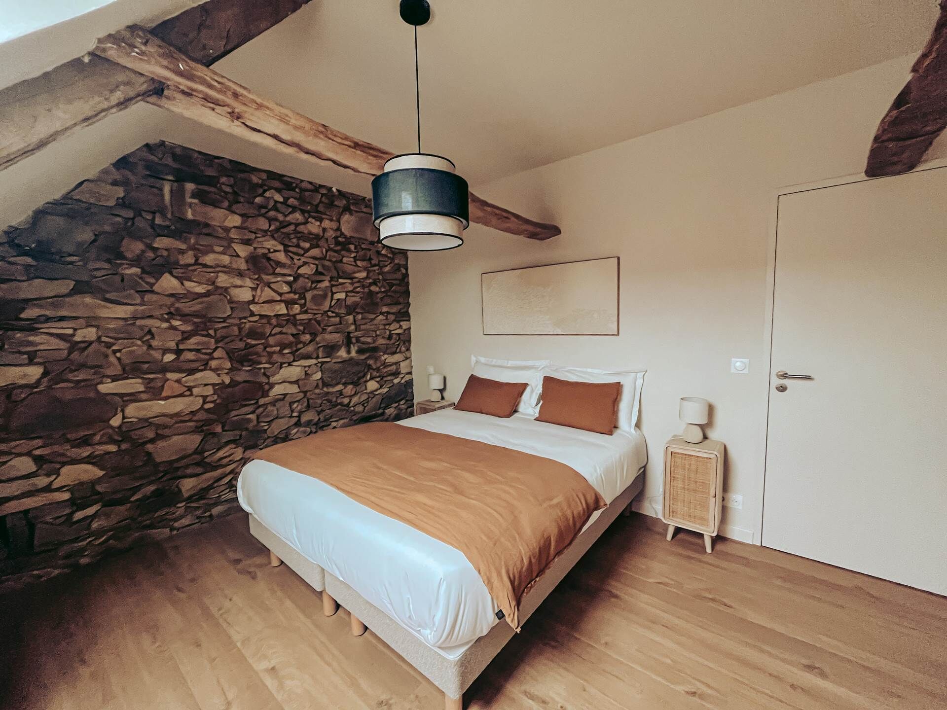 A bedroom with a stone wall, wooden floor, a bed with a brown blanket, a bedside table, and a hanging light fixture.