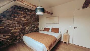 A bedroom with a stone wall, wooden floor, a bed with a brown blanket, a bedside table, and a hanging light fixture.