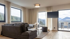 A modern living room with a grey sectional sofa, a small round table, and a flat-screen TV. Large windows offer a view of a mountainous landscape. - Penthouse for up to 8 people & infinity pool (St. Martin am Tennengebirge)