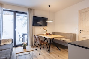 A modern living room with a sofa, a dining table with chairs, a TV mounted on the wall, and a wooden floor.