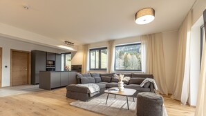 A modern living room with a sofa, ottoman, and coffee table, a kitchen area, and large windows with curtains.