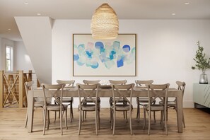 A dining area with a wooden table and chairs, a woven pendant light, and a framed abstract painting on the wall.