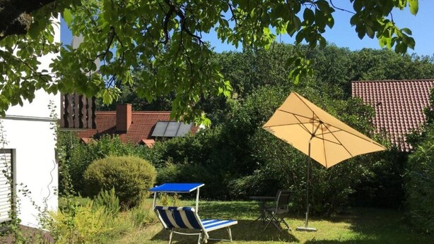 Un jardín con césped, una silla azul y blanca a rayas, un paraguas amarillo y un panel solar en una casa.