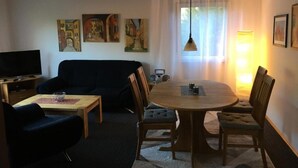 Una sala de estar con un sofá de color oscuro, una mesa de comedor de madera con cuatro sillas, un televisor sobre un mueble y obras de arte en las paredes.