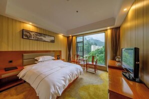 A hotel room with a large bed, a desk with a TV, and a window offering a view of greenery and buildings. - Leshanshui Holiday Hotel (Yizhang)