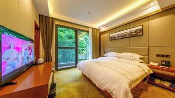 A hotel room with a bed, a TV, a desk, a nightstand, and a window with a view of greenery.