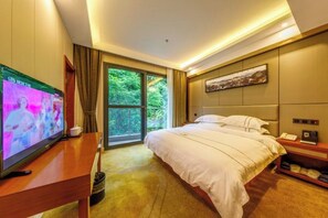 A hotel room with a bed, a TV, a desk, a nightstand, and a window with a view of greenery.