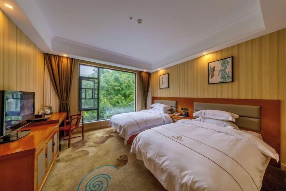 A hotel room with two beds, a desk with a TV, a window with a view of greenery, and a painting on the wall.