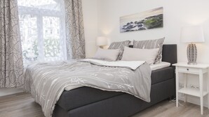Quarto com cama, mesa de cabeceira, luminária, pintura e janela com cortinas.