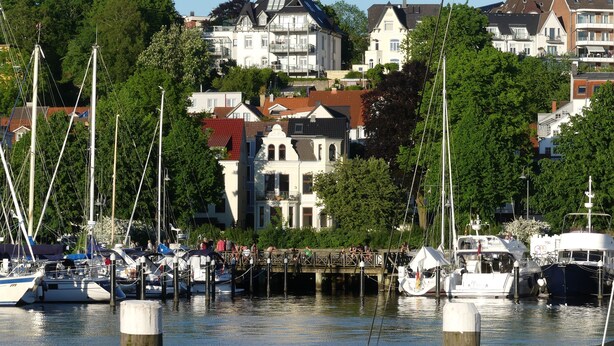 Uma marina com barcos atracados, um cais de madeira e edifícios ao fundo.