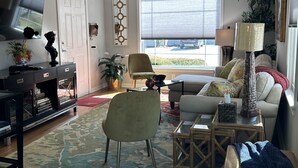 Una sala de estar con sofá, sillones, una mesa de centro, televisor, estantería y ventana con persianas.