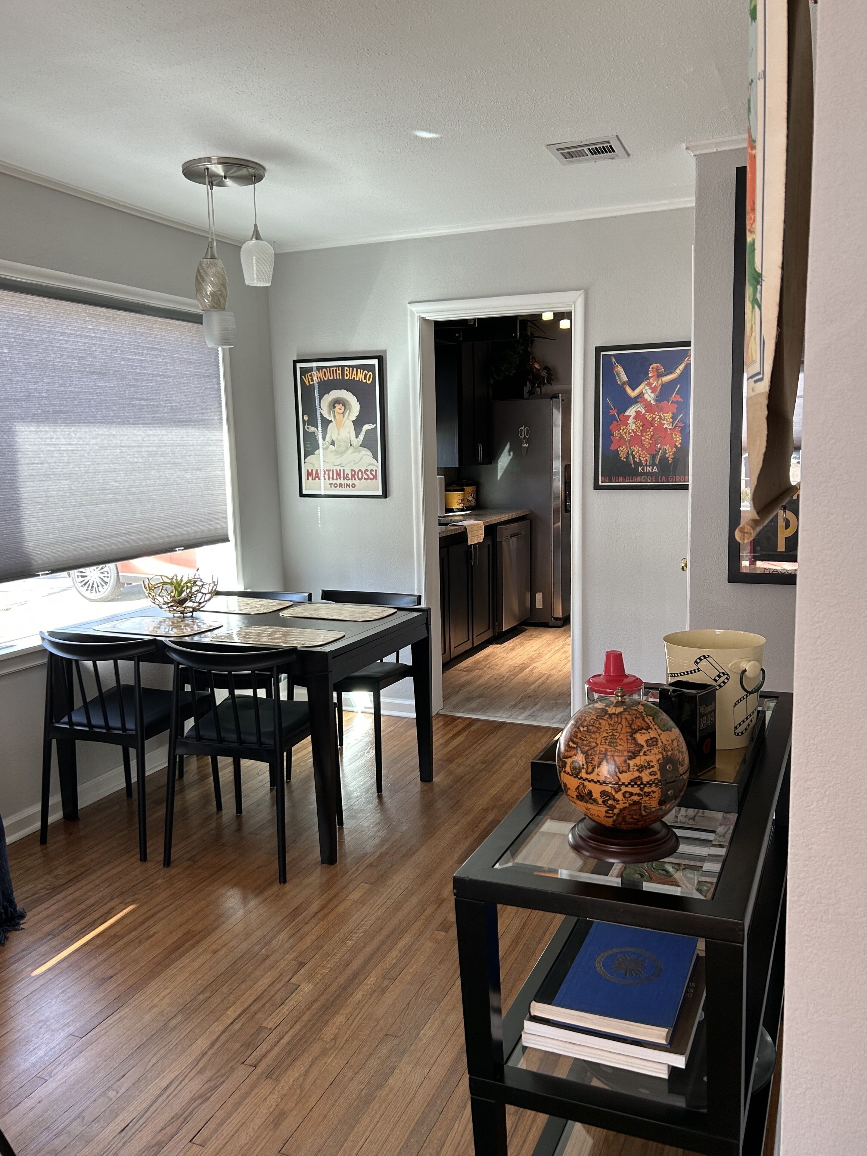 A dining area with a table and chairs, a kitchen in the background, and a poster on the wall.
