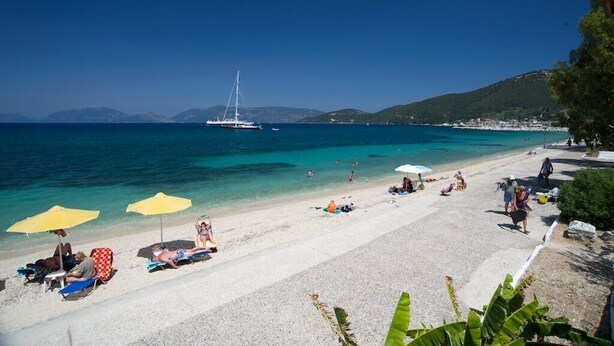 Een strand met helder turquoise water, een zeilboot in de verte en mensen die zonnebaden.