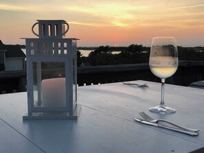 Une table avec un verre de vin blanc, une lanterne et des couverts, sur un balcon donnant sur un coucher de soleil.