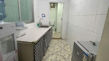 Un baño pequeño y estrecho con paredes y suelo de azulejos blancos, un lavamanos, un espejo y un dispensador de agua.