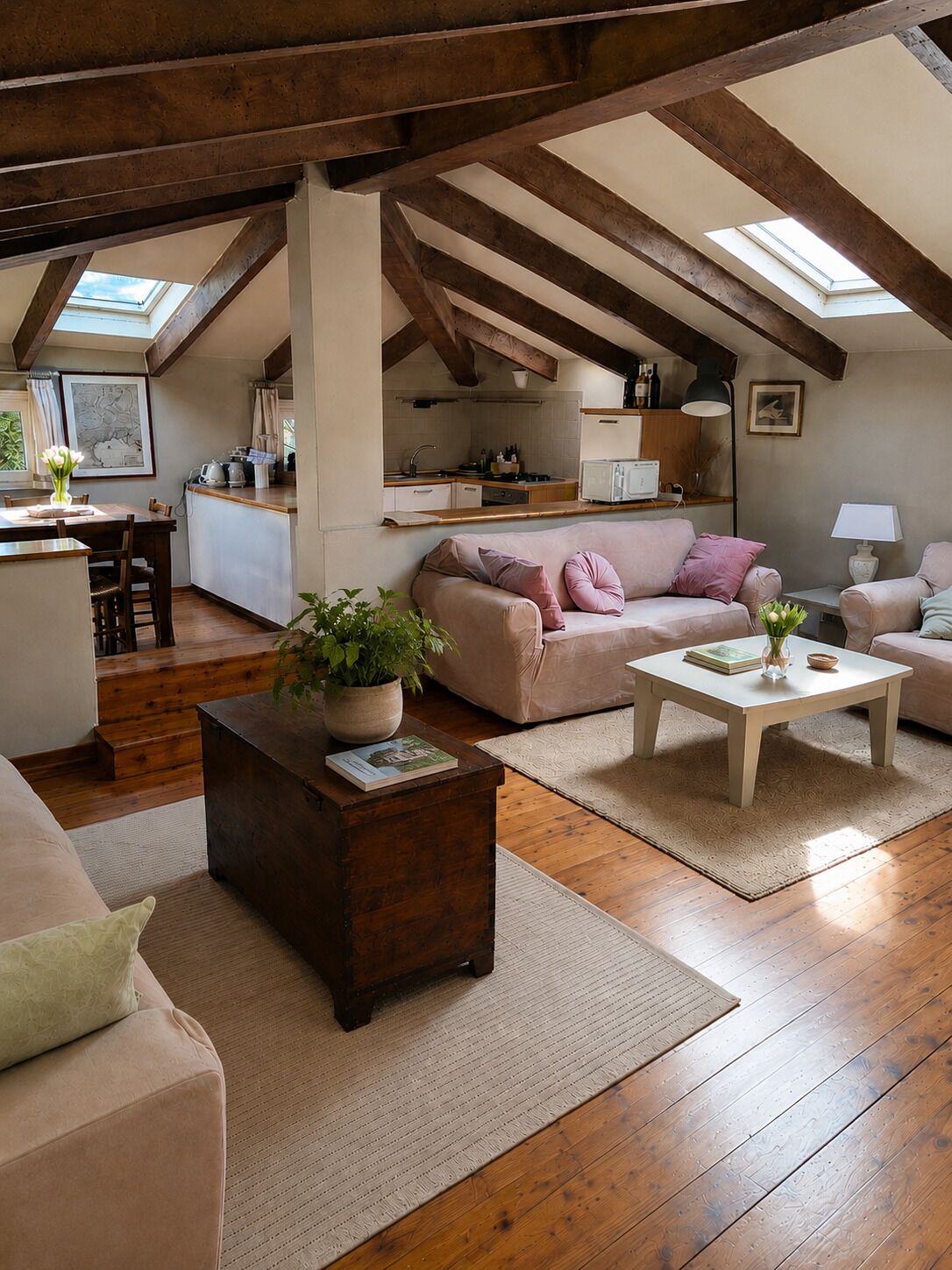 A spacious loft with a high ceiling, exposed wooden beams, a kitchen area, a seating area with a sofa and armchairs, and a dining table.
