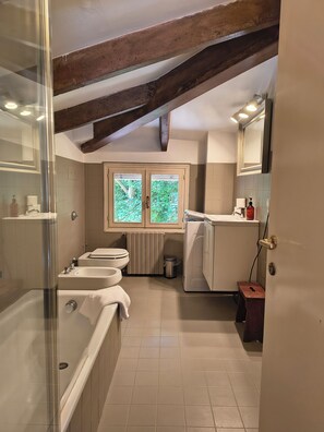 A bathroom with a bathtub, two sinks, a toilet, a radiator, and a window with a view of greenery. - Your getaway to discover the Veneto. Only 5 Minutes from central Padova (Sant'Agostino)