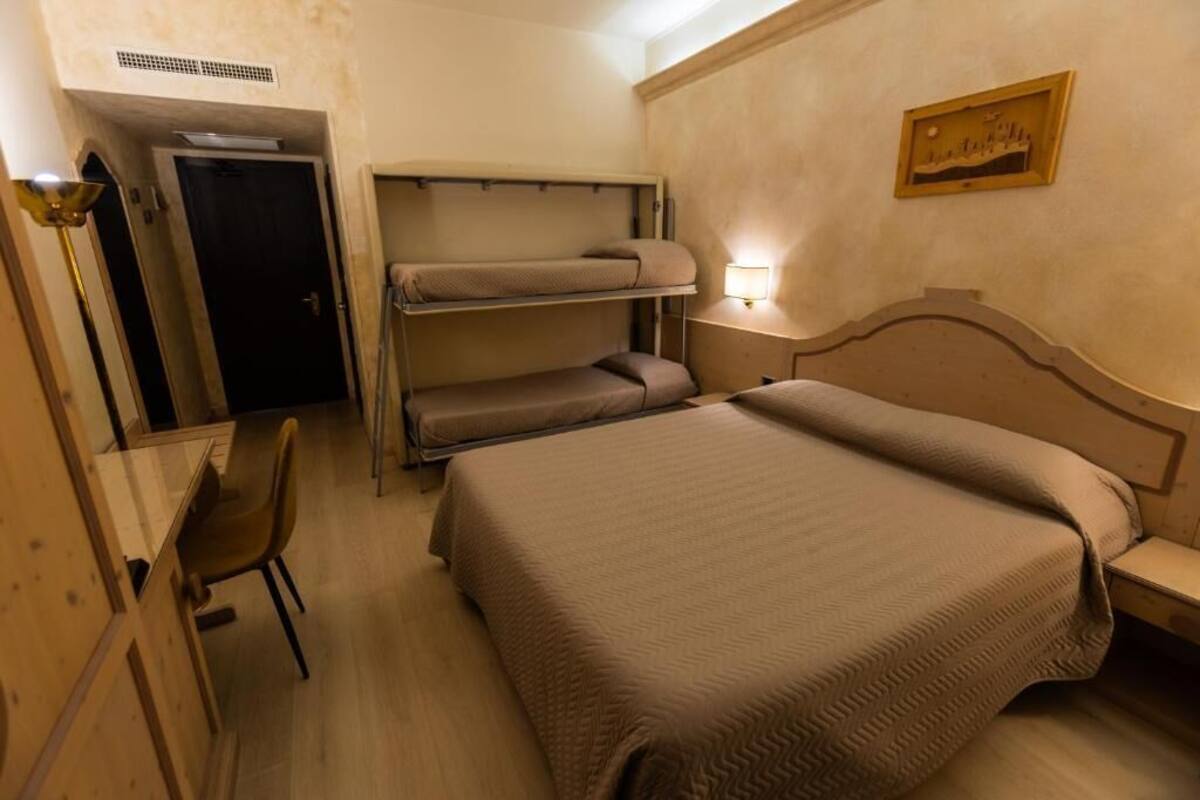 A hotel room with a bunk bed, a desk with a chair, a bedside table, and a wall-mounted lamp.