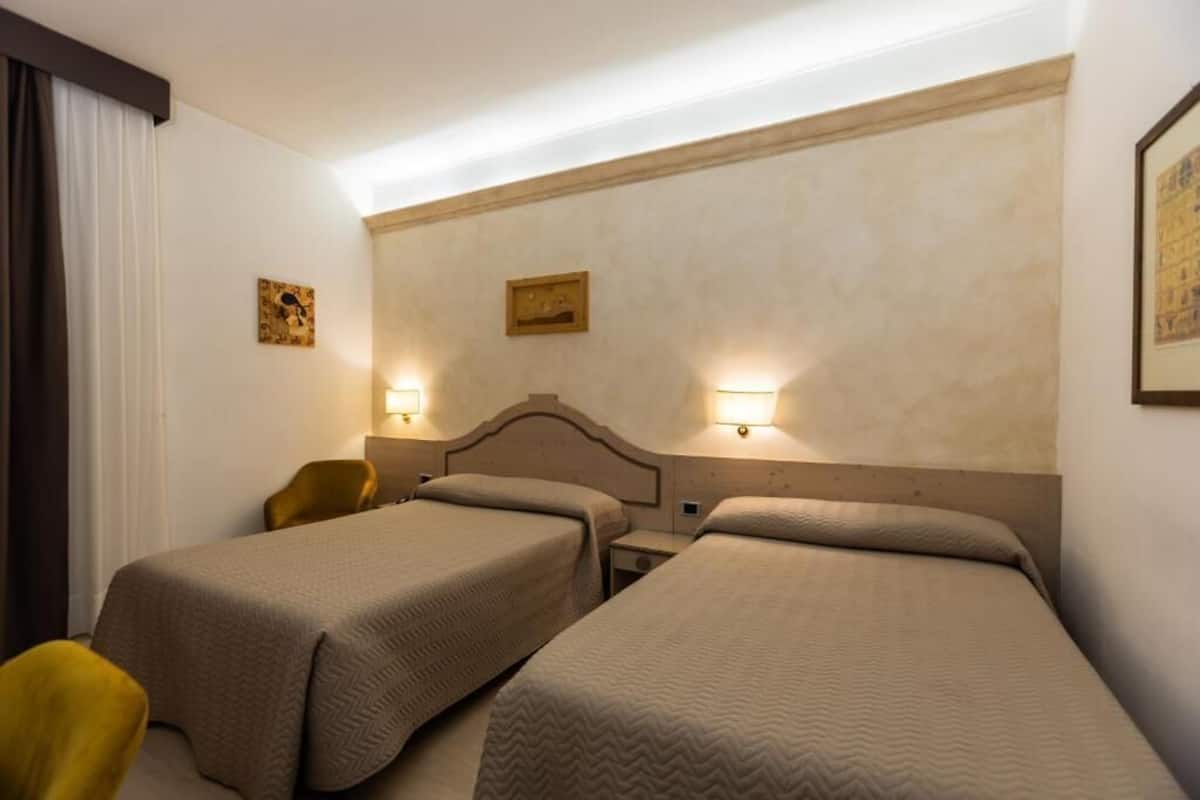 A hotel room with two single beds, a nightstand, a chair, and wall-mounted lamps.