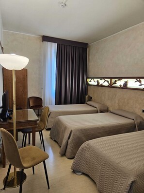 Quarto de hotel com duas camas, uma escrivaninha, uma cadeira, um abajur e uma janela com cortinas.