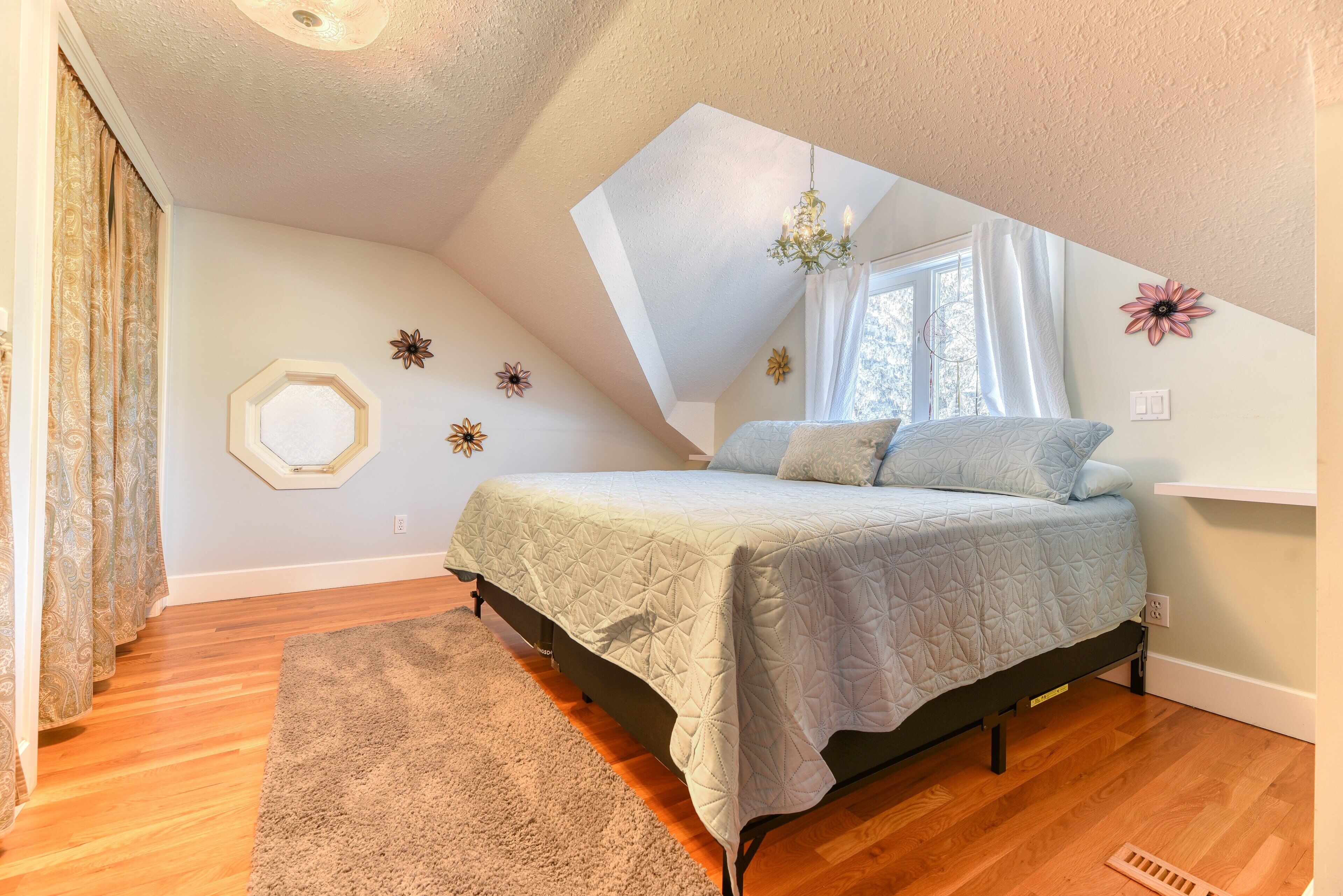 Une chambre avec un plafond en pente, un lit recouvert d’une couvre-lit à motifs géométriques, un lustre et une fenêtre avec des rideaux.