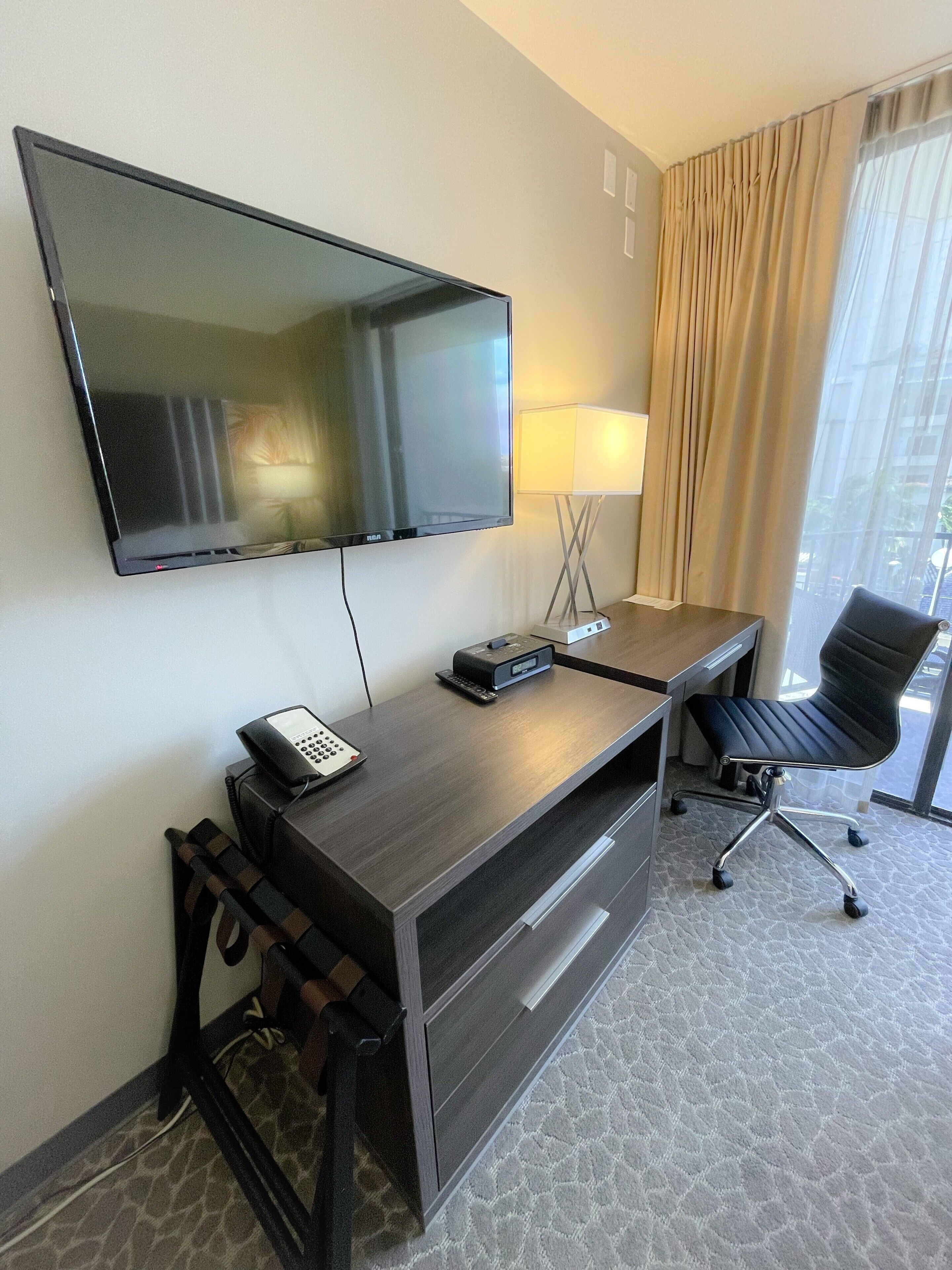 A hotel room with a flat-screen TV mounted on the wall, a desk with a telephone, a lamp, and a chair.