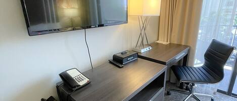 A hotel room with a flat-screen TV mounted on the wall, a desk with a telephone, a lamp, and a chair.