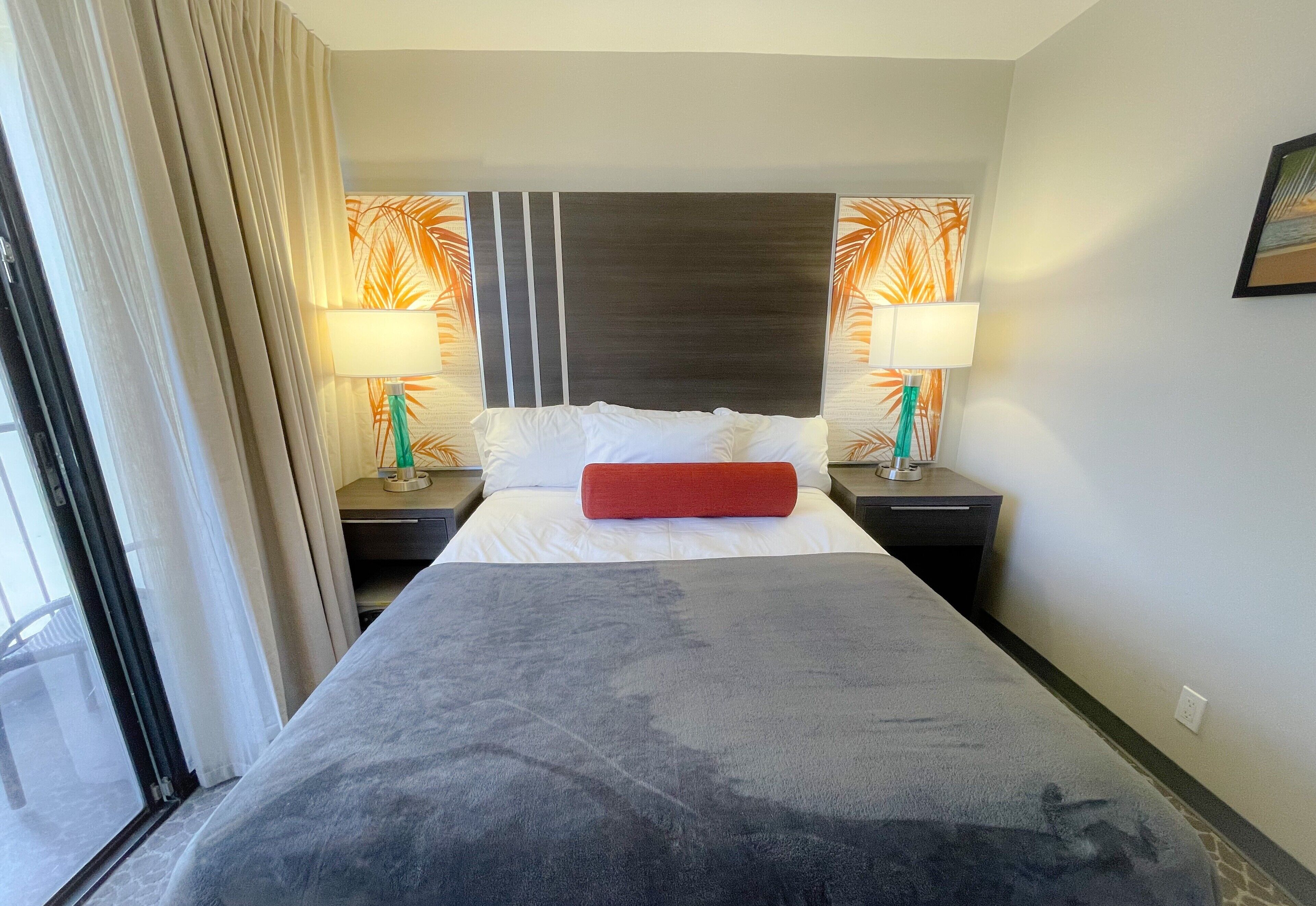 Een hotelkamer met een groot bed, twee nachtkastjes met lampen, een hoofdbord met een bladmotief en een raam met gordijnen.