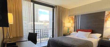 Hotelkamer met een bed, bureau, stoel, televisie, lamp en uitzicht op de stad door grote ramen.
