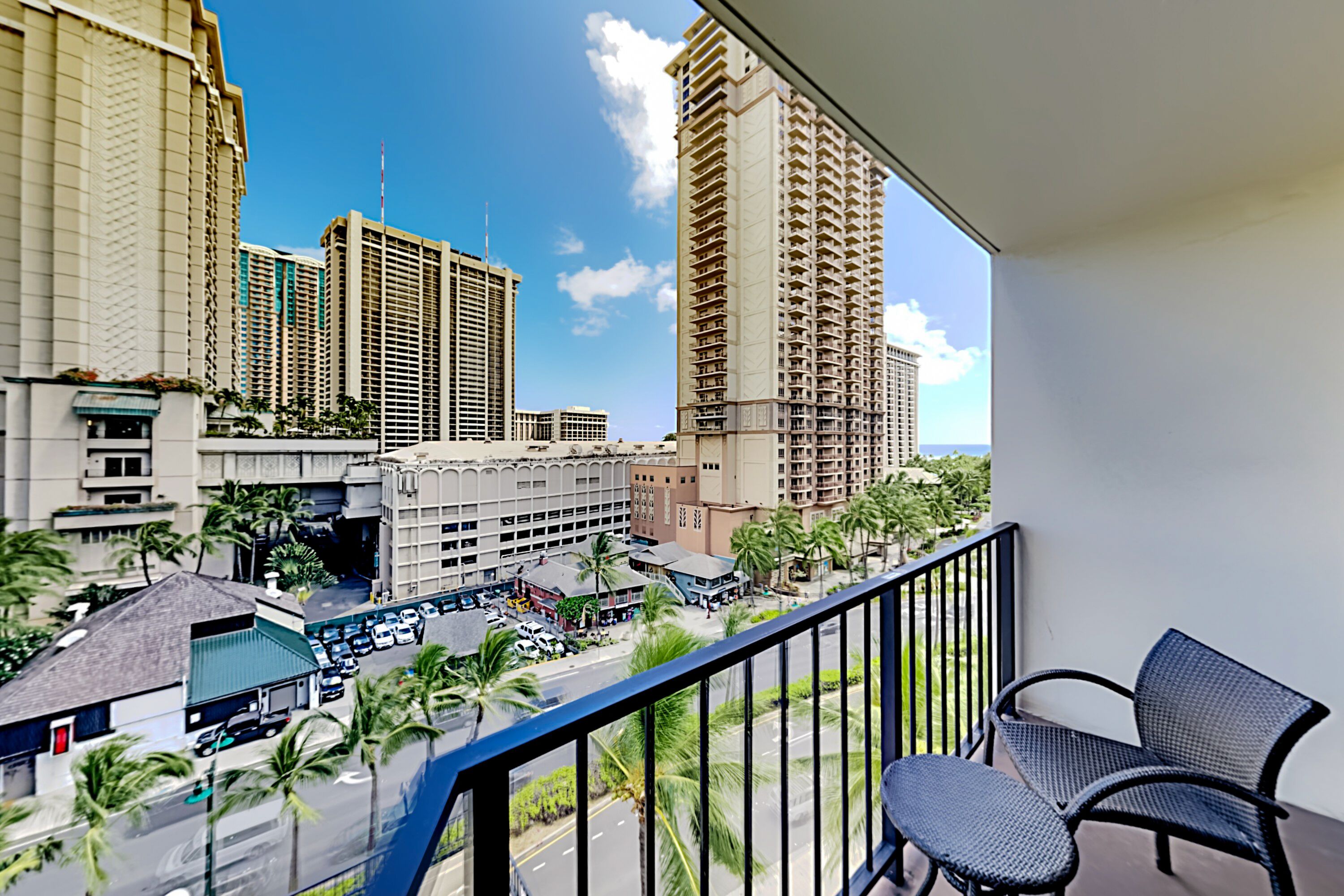 A balcony with a view of tall buildings, a road with cars, and palm trees.
