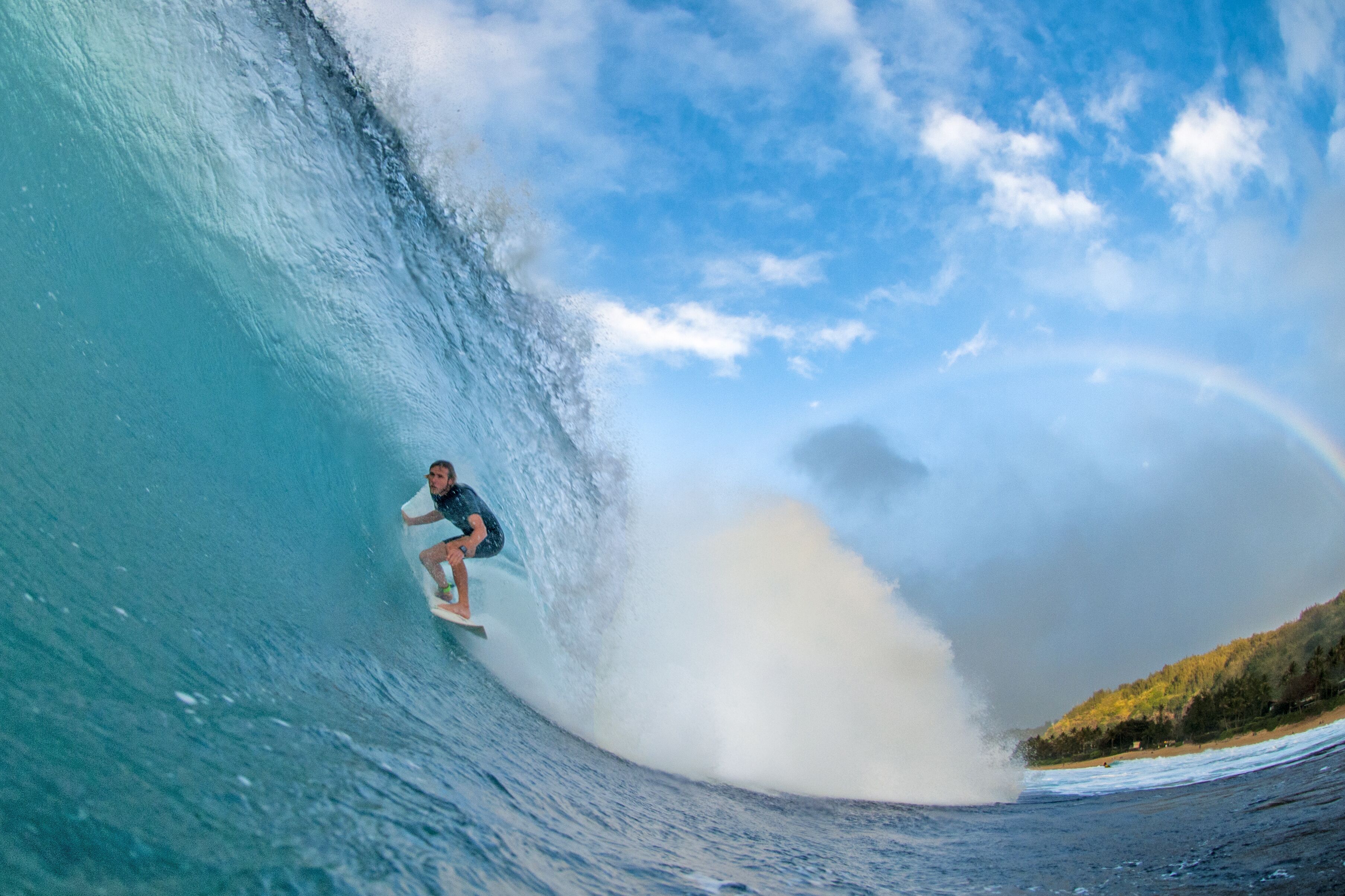 Un surfeur chevauchant une grosse vague, avec un ciel dégagé et un arc-en-ciel en arrière-plan.