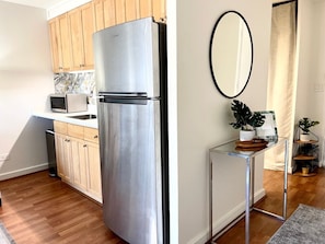Cocina compacta con armarios de madera, un refrigerador de acero inoxidable, un microondas y una pequeña mesa con un espejo y plantas.