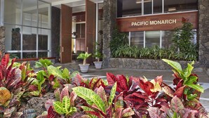 Entrada de hotel con un letrero que dice "PACIFIC MONARCH", rodeada de exuberante follaje y plantas en macetas.