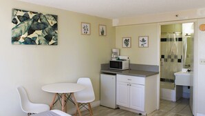 Una cucina funzionale con frigorifero bianco, forno a microonde e macchina da caffè sul piano di lavoro. C'è un piccolo angolo ristoro con due sedie bianche e un tavolo rotondo. Le pareti sono decorate con stampe botaniche incorniciate.