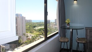Ein Zimmer mit einem großen Fenster, das einen Blick auf Stadt und Grünflächen bietet. Auf der Fensterbank stehen ein kleiner Tisch, zwei Stühle und ein Blumentopf.